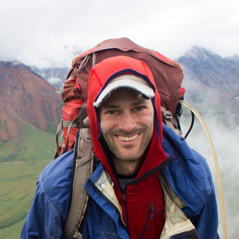 A man with a backpack smiles at the camera. He is wearing a blue jacket and a red hooded sweatshirt. He also has a baseball cap on. He appears to be in a mountainous area, with mountains and fog visible in the background. He seems to be on an outdoor adventure.
